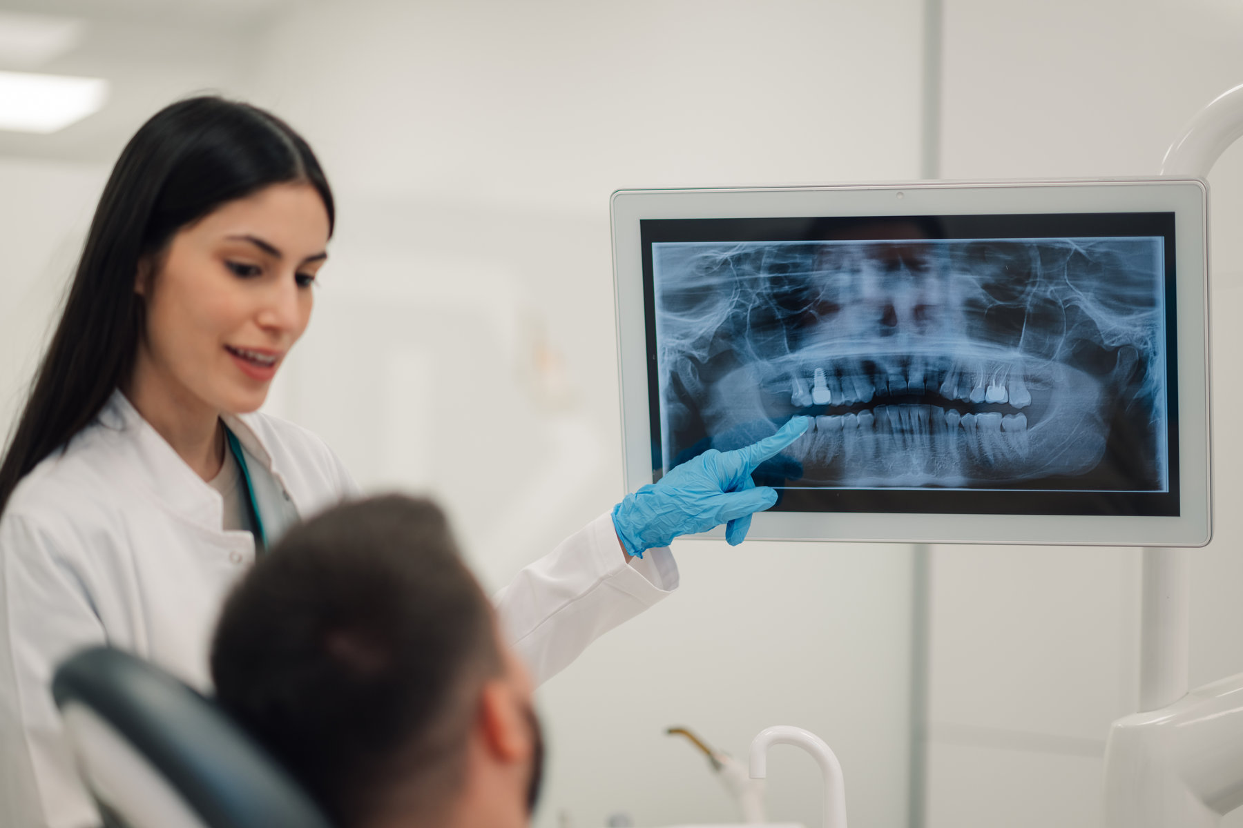 Female dentist wearing blue gloves pointing at digital screen showing teeth radiography to patient in dental clinic, explaining diagnosis and treatment