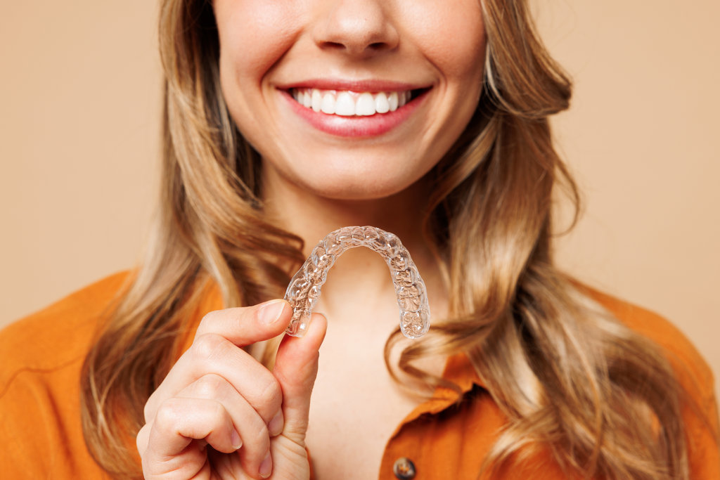 Close up cropped young happy woman wear orange shirt casual clothes hold in hand invisible transparent aligners, invisalign bracer isolated on plain pastel light beige background. Lifestyle concept