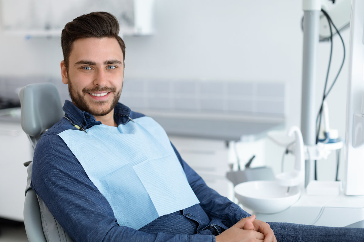 Side view of handsome young man patient sitting in dental chair and smiling, panorama with copy space. Cheerful millennial man showing beautiful white smile after treatment at modern dental clinic