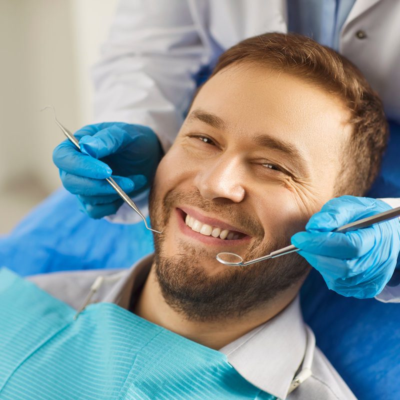 Dental health. Man sits comfortably in dentists chair, showing off white smile that exudes confidence. Dentist holds special tools in his hands for preventive and therapeutic examination of teeth.